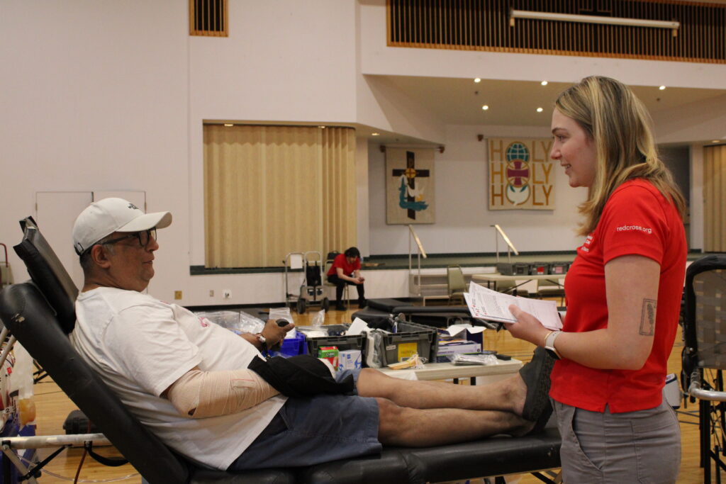 Man lies on a blood donor bed as he answers questions with Red Cross reporter.